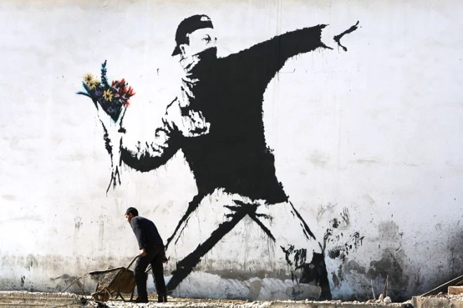 BETHLEHEM, WEST BANK - DECEMBER 05:  A Palestinian labourer works under a large wall painting by elusive British graffiti artist Banksy December 5, 2007 on a building wall in the biblical city of Bethlehem in the West Bank. The Bristol-born artist has adorned Israel&rsquo;s West Bank separation barrier and Bethlehem walls with new images, including one of a dove wearing a flak jacket and a soldier being frisked by a young girl. His works, along with those of other international artists, are part of an exhibition called Santa&rsquo;s Ghetto.  (Photo by David Silverman/Getty Images)